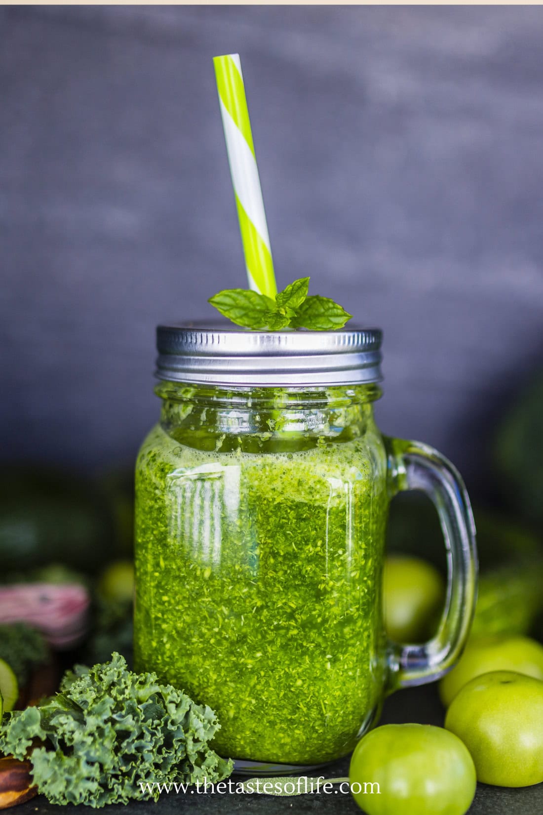 A mason jar filled with a bright green smoothie, topped with mint leaves and a green-striped straw, surrounded by fresh green vegetables and fruits.