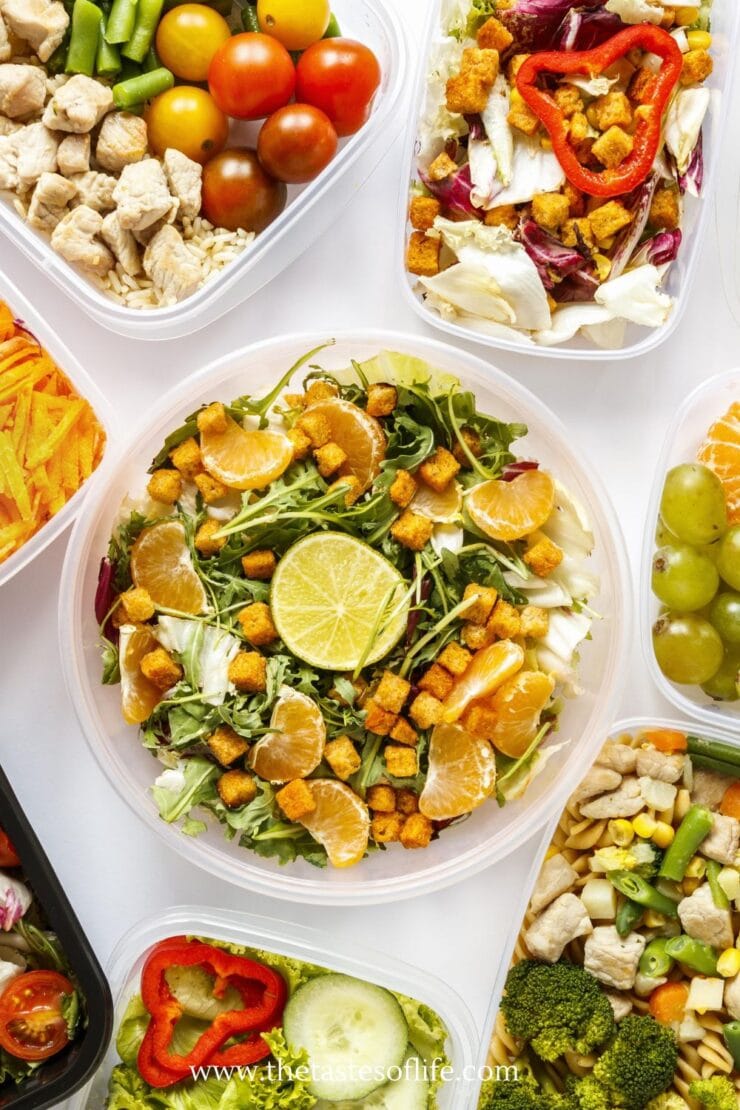 A variety of meal prep containers filled with colorful salads, fruits, vegetables, cooked chicken, croutons, and garnished with lime and orange slices, arranged on a white surface.