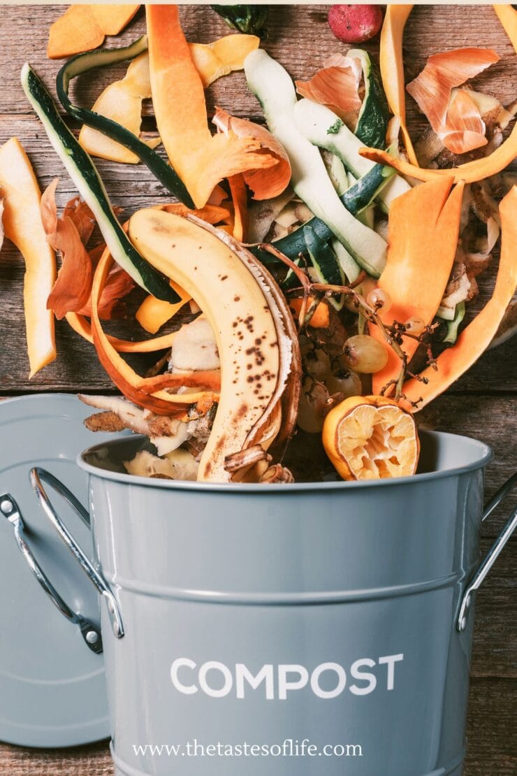 A gray compost bin filled with assorted fruit and vegetable scraps, including a banana peel, citrus, carrot, and zucchini peels, sitting on a wooden surface. The word “COMPOST” is printed on the bin.