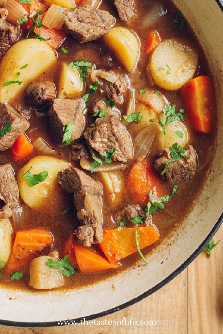 A pot of beef stew with chunks of beef, potatoes, carrots, onions, and fresh parsley in a rich brown broth.