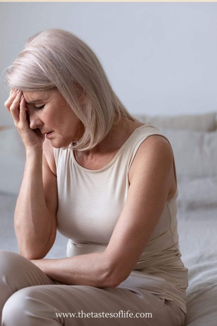 A woman with light hair sits on a bed, looking distressed and holding her head in her hand as if experiencing pain or sadness. She is wearing a sleeveless top. The background is softly lit and neutral.