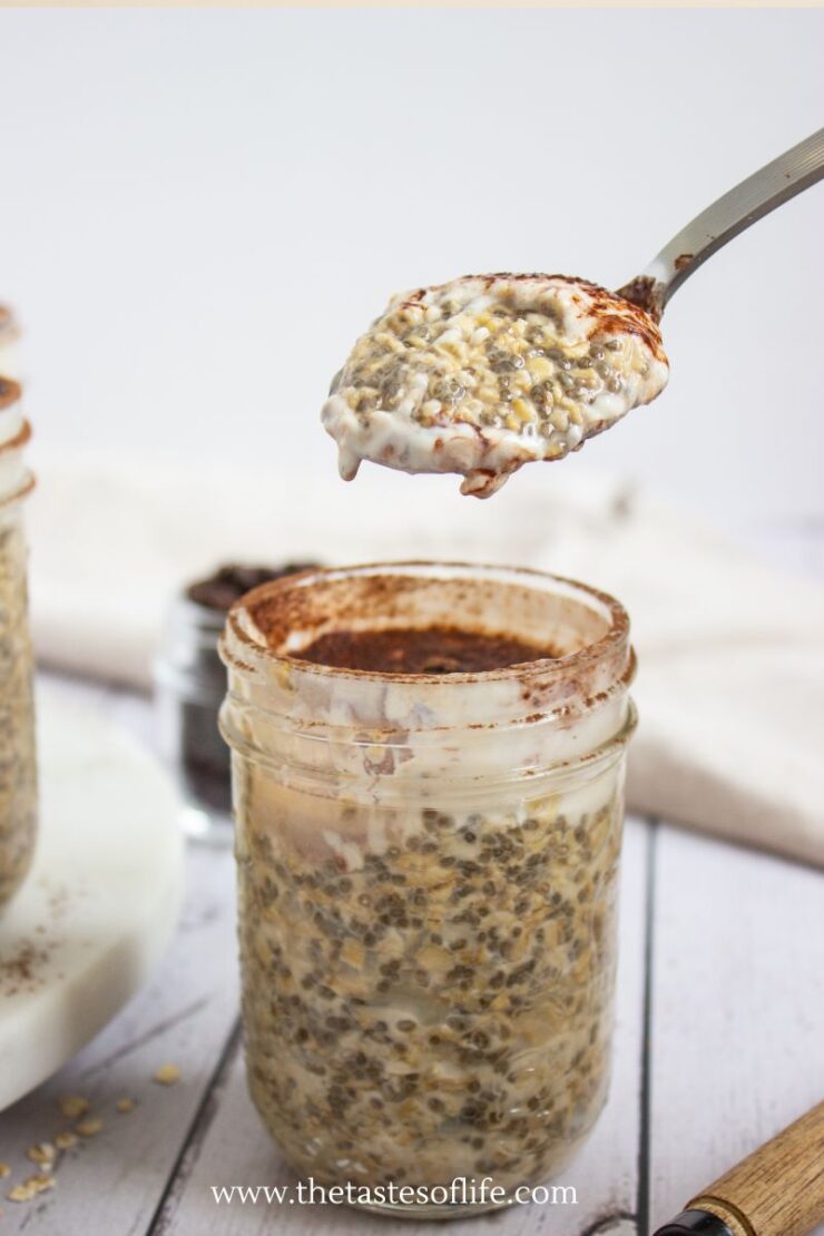 A spoonful of chia pudding being lifted from a glass jar, with creamy layers and sprinkled with cocoa powder. The jar sits on a light-colored surface, and the dessert appears thick and textured.