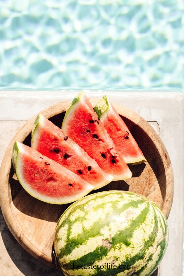 A wooden plate with watermelon slices and a whole watermelon sits by a swimming pool, with clear blue water in the background.