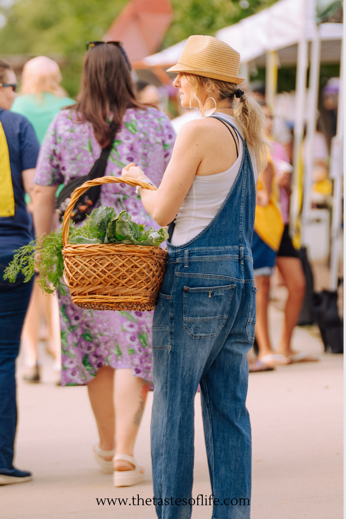 A person wearing overalls and a straw hat holds a basket of leafy greens at an outdoor market, surrounded by people walking and shopping.