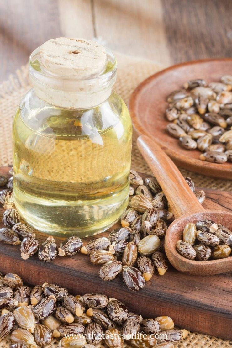 A glass bottle of castor oil with a cork stopper surrounded by castor beans on a wooden board, with a wooden spoon and a small bowl of beans nearby to help explain 8 ways castor oil balance hormones.