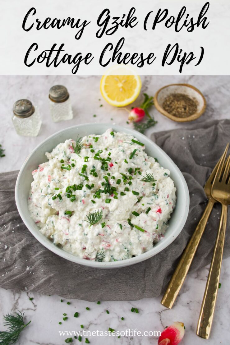 A bowl of creamy Polish cottage cheese dip (Gzik) topped with chopped chives and dill, next to gold utensils, pepper, salt shakers, lemon, and herbs, on a marble surface with a gray napkin.