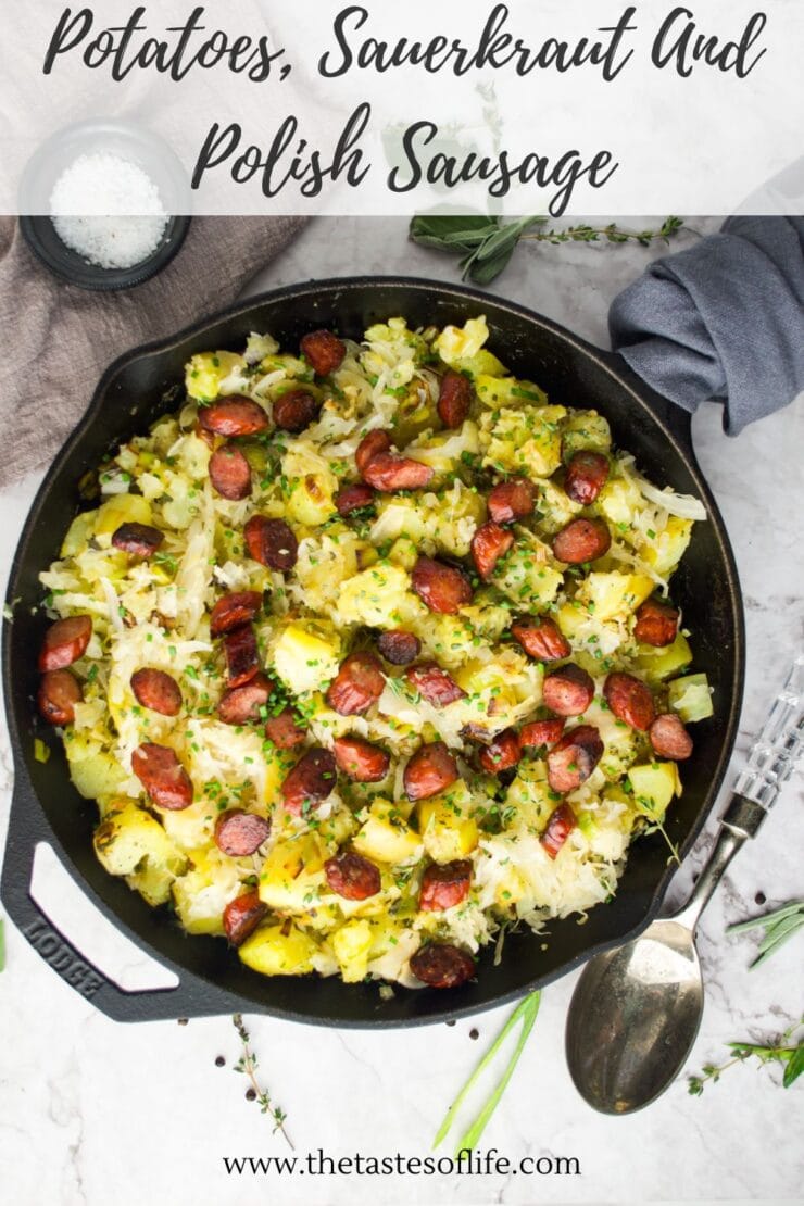 A skillet filled with sliced potatoes, sauerkraut, and pieces of Polish sausage garnished with herbs, on a marble surface with a spoon, a gray napkin, and a small bowl of salt nearby.