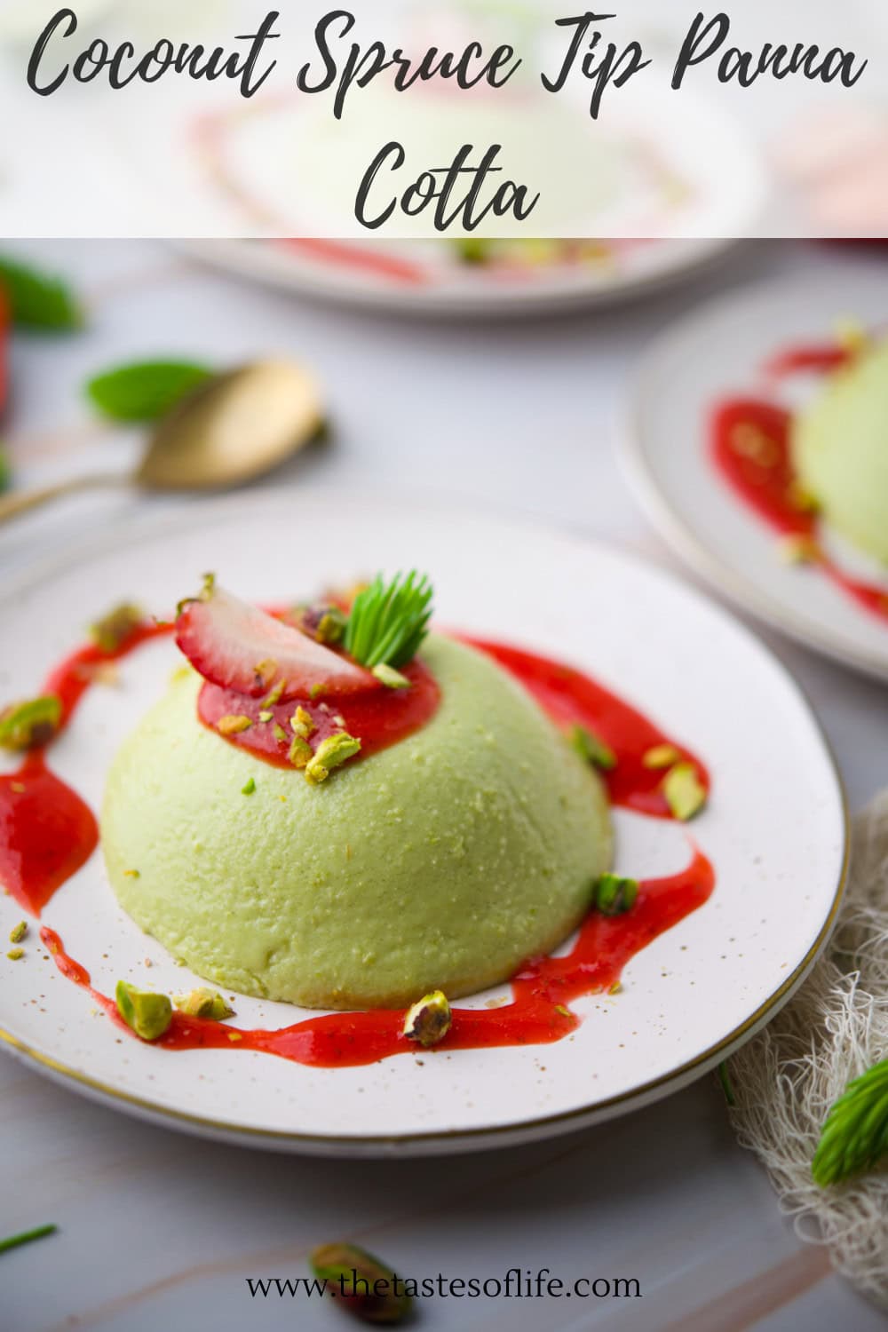 A dome-shaped green panna cotta sits on a white plate with raspberry sauce, chopped pistachios, a strawberry slice, and a small spruce tip garnish. A gold spoon and another plate are visible in the background.