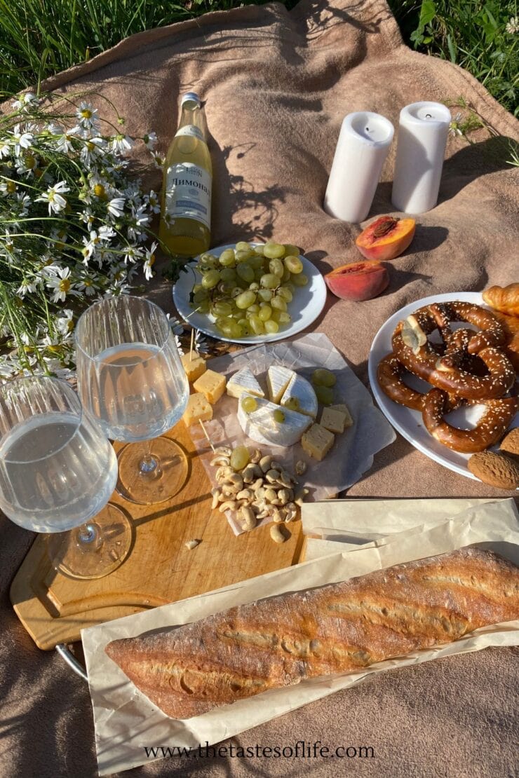 A picnic setup on a blanket with bread, cheese, grapes, pretzels, peach halves, wine, two glasses, candles, and a bouquet of wildflowers, all arranged outdoors in the sunlight.