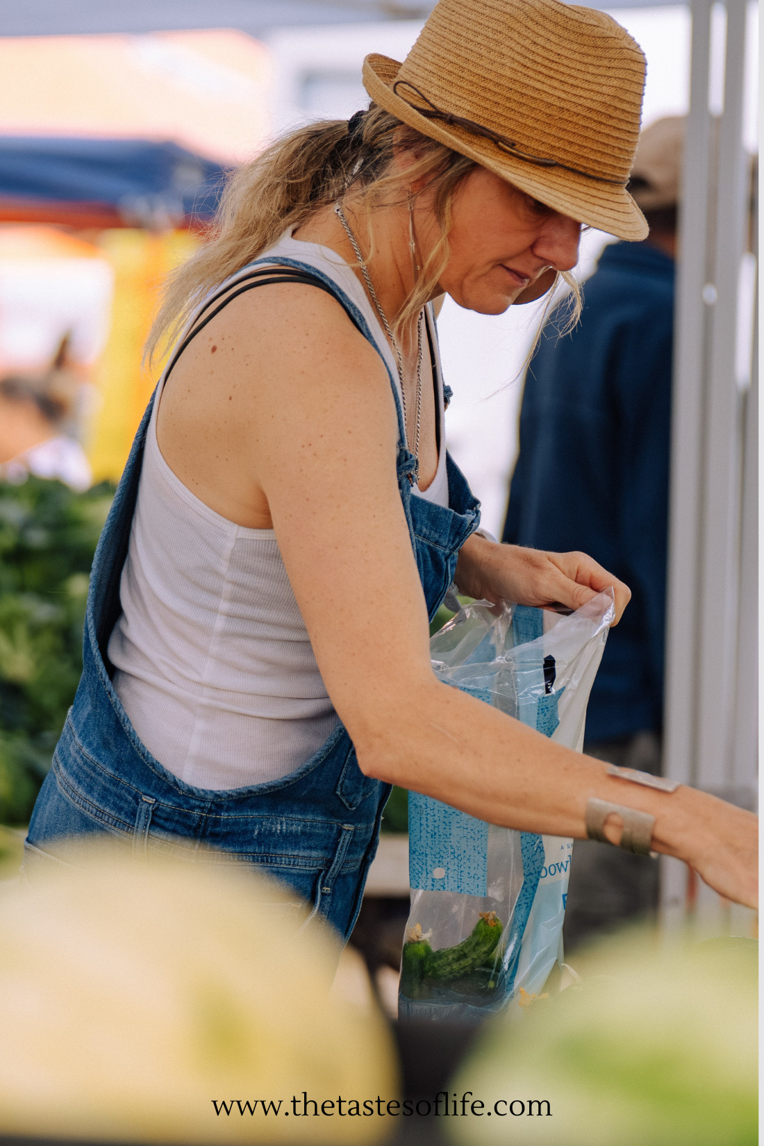A woman in denim overalls and a straw hat shops at an outdoor market, placing produce into a plastic bag. Fresh greens and other vegetables are visible around her.