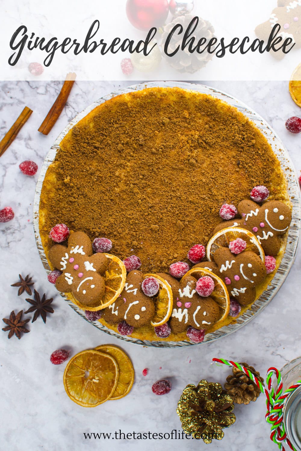 A round gingerbread cheesecake is topped with gingerbread cookies, candied cranberries, dried orange slices, and dusted crumbs, set on a marble surface with festive holiday decorations around it.