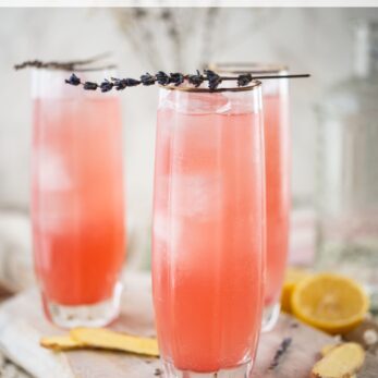 Three tall glasses filled with a pink rhubarb, lavender, and hibiscus cooler, garnished with lavender sprigs. Slices of lemon and ginger are on the wooden surface. Recipe title is displayed at the top.