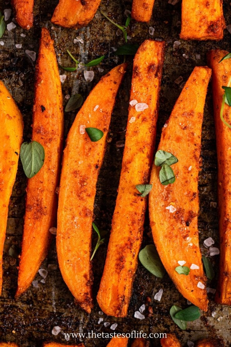 Close-up of roasted sweet potato wedges sprinkled with coarse salt and fresh herb leaves on a baking tray.