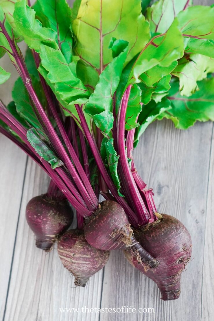 Four fresh beetroots with leafy green tops rest on a wooden surface. The beets have deep red-purple bulbs and long stems, with bright green leaves above.