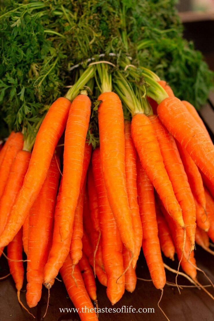 A bunch of fresh, bright orange carrots with green tops, arranged closely together on a dark surface. The roots of the carrots are visible, and they look freshly harvested.