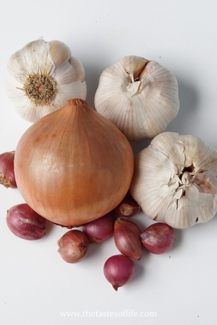 A large brown onion, three bulbs of garlic, and seven small red shallots are arranged on a white surface.