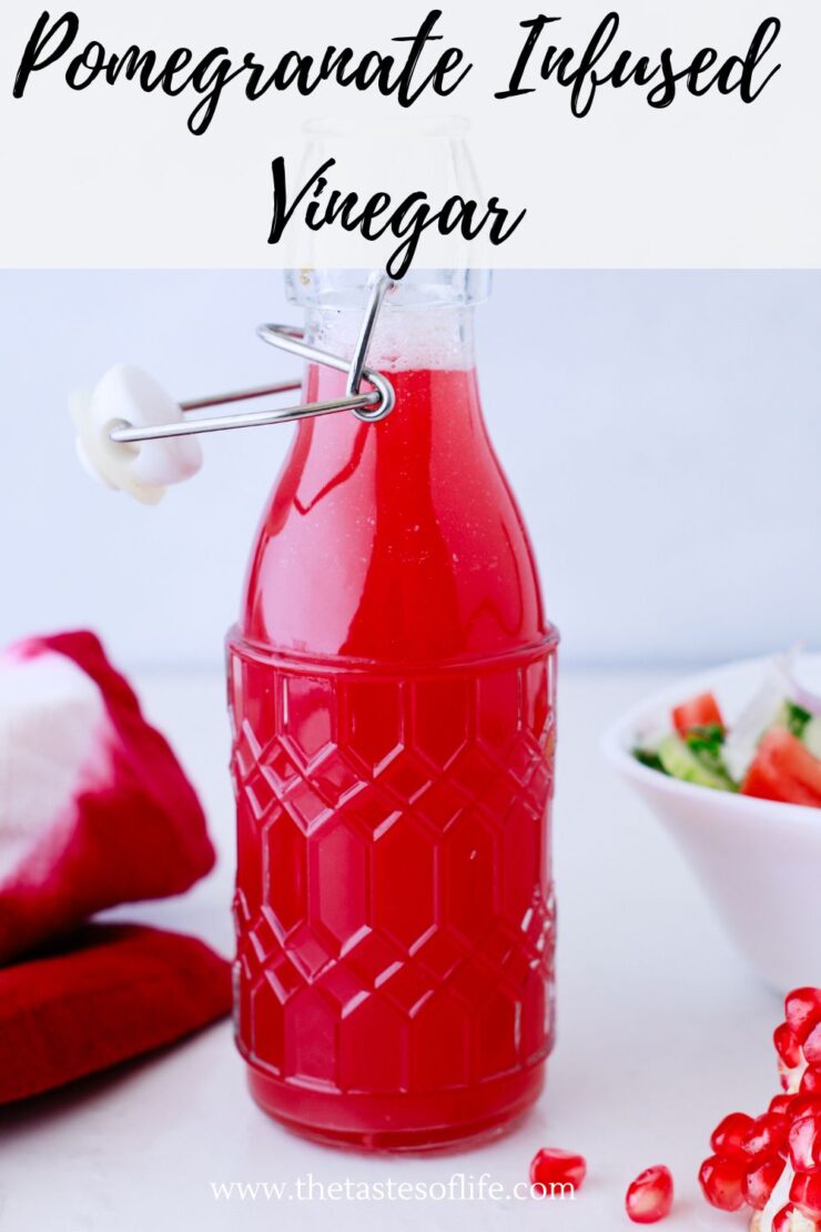 A glass bottle filled with bright red pomegranate infused vinegar sits on a white surface, with a bowl of salad, pomegranate seeds, and a red cloth nearby. The bottle has a decorative pattern and a swing top lid.