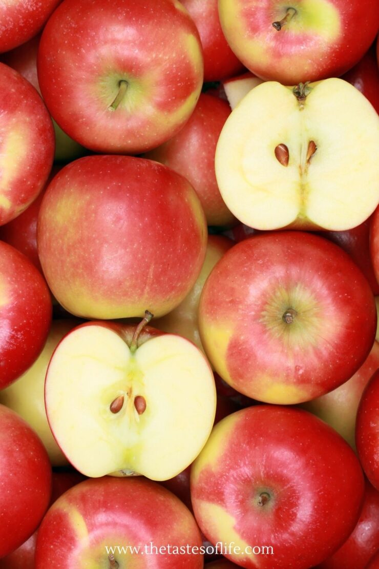 A pile of red apples, with some cut in half to show the yellowish flesh and seeds inside. The texture and color variations are visible on each apple.