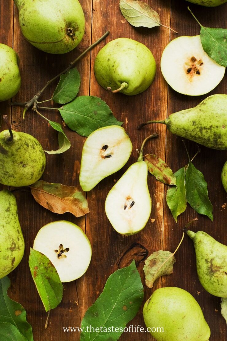 Several whole and sliced green pears with leaves are scattered on a rustic wooden table. Some pears are halved, revealing seeds. Text at the bottom reads www.thetastesoflife.com..