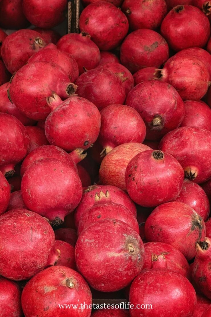 A pile of fresh, ripe pomegranates with smooth, red skins fills the frame; one fruit in the middle is slightly lighter in color than the others.