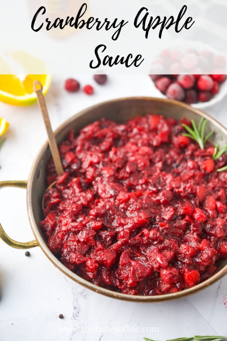 A rustic bowl filled with chunky cranberry apple sauce, garnished with a sprig of rosemary. A spoon rests in the bowl, and fresh cranberries, lemon, and herbs are scattered around. Text reads Cranberry Apple Sauce.