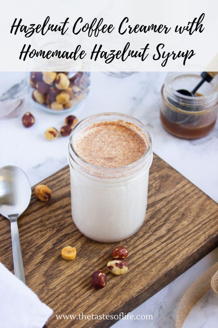 A glass jar filled with hazelnut coffee creamer, topped with cinnamon, sits on a wooden board with scattered hazelnuts. A spoon, a jar of syrup, and a bowl of hazelnuts are nearby. Text above reads, Hazelnut Coffee Creamer with Homemade Hazelnut Syrup.