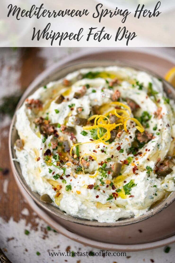 A bowl of Mediterranean spring herb whipped feta dip, garnished with chopped herbs, olive oil, lemon zest, and red pepper flakes, sits on a wooden table. The dip looks creamy and fresh.