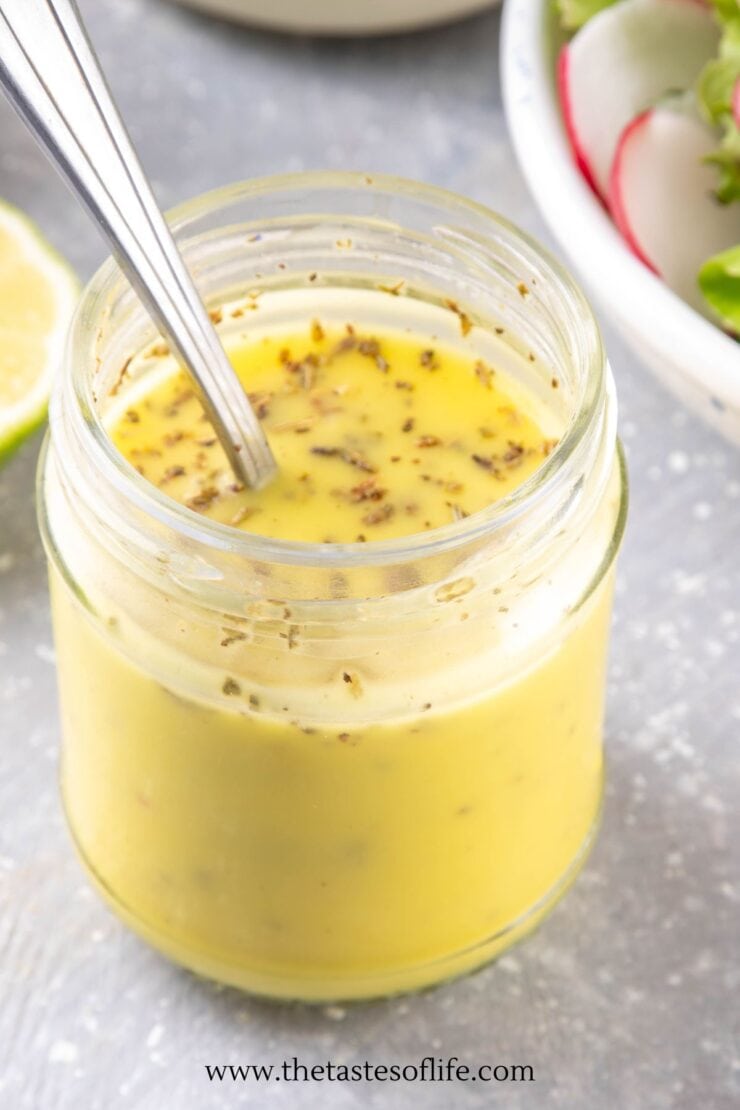 A glass jar filled with yellow salad dressing, topped with herbs, and a spoon inside. A salad and a lemon wedge are partially visible beside the jar.