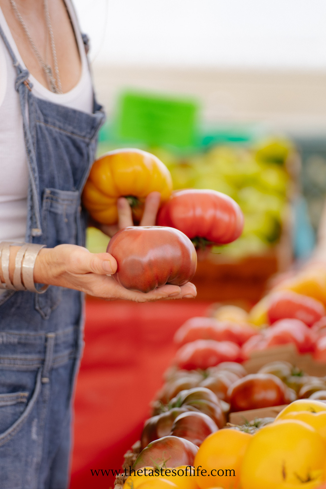 A person in denim overalls holds a large heirloom tomato at a market stand filled with colorful tomatoes. The background is blurred, highlighting the produce.