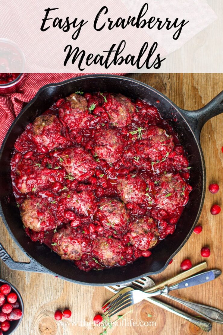 A cast iron skillet filled with meatballs covered in a chunky cranberry sauce, garnished with herbs. Fresh cranberries and utensils are scattered on a wooden table around the skillet. Text at the top reads Easy Cranberry Meatballs.
