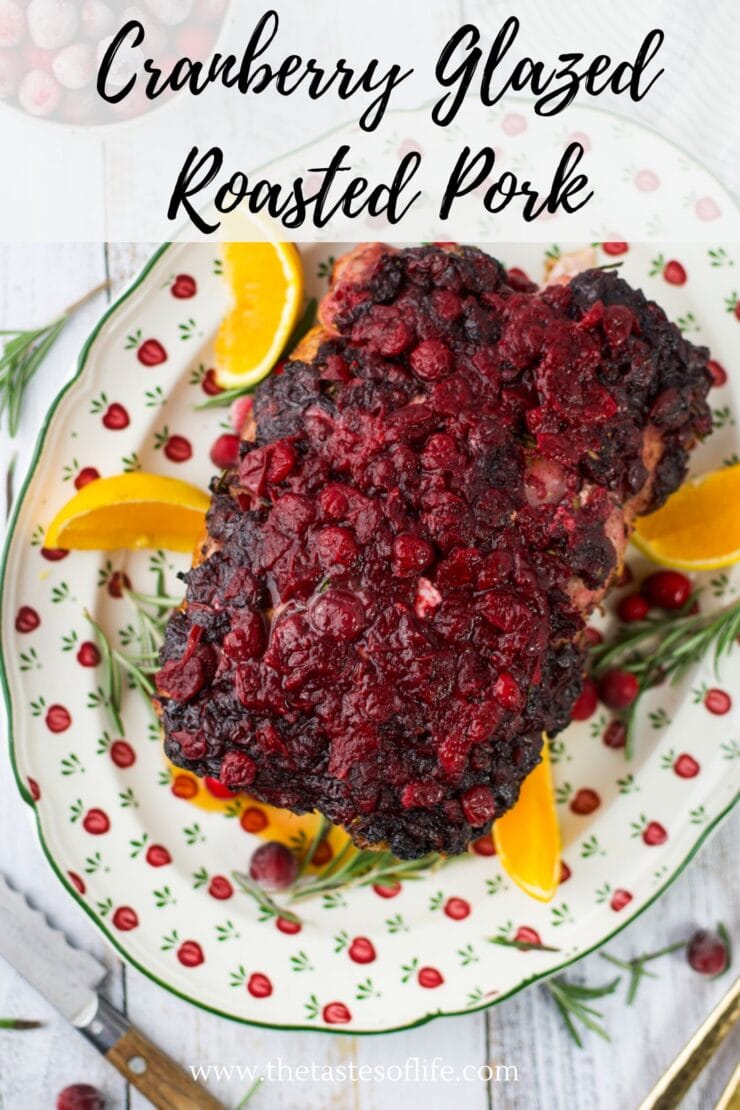 Overhead view of cranberry glazed roasted pork on a festive plate, garnished with orange slices, fresh cranberries, and rosemary sprigs. The dish is placed on a white tablecloth. Text reads Cranberry Glazed Roasted Pork.