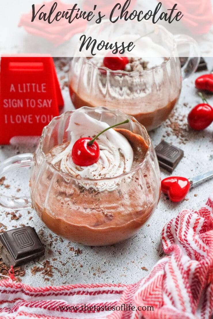 A glass cup of chocolate mousse topped with whipped cream and a cherry, surrounded by chocolate pieces, cherries, a red heart, and a sign reading A little sign to say I love you. Red and white cloth is nearby.