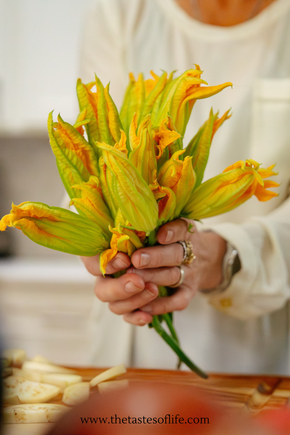 A person holds a fresh bouquet of yellow zucchini blossoms in both hands, with a blurred kitchen background. The website www.thetastesoflife.com is visible at the bottom of the image.