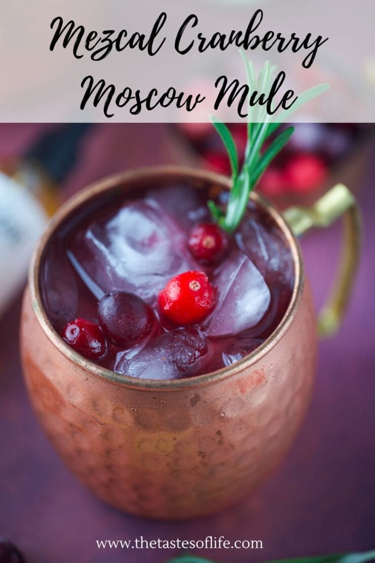 A copper mug filled with ice, cranberries, and a rosemary sprig, containing a Mezcal Cranberry Moscow Mule cocktail. Text above reads Mezcal Cranberry Moscow Mule, with the website thetastesoflife.com below.