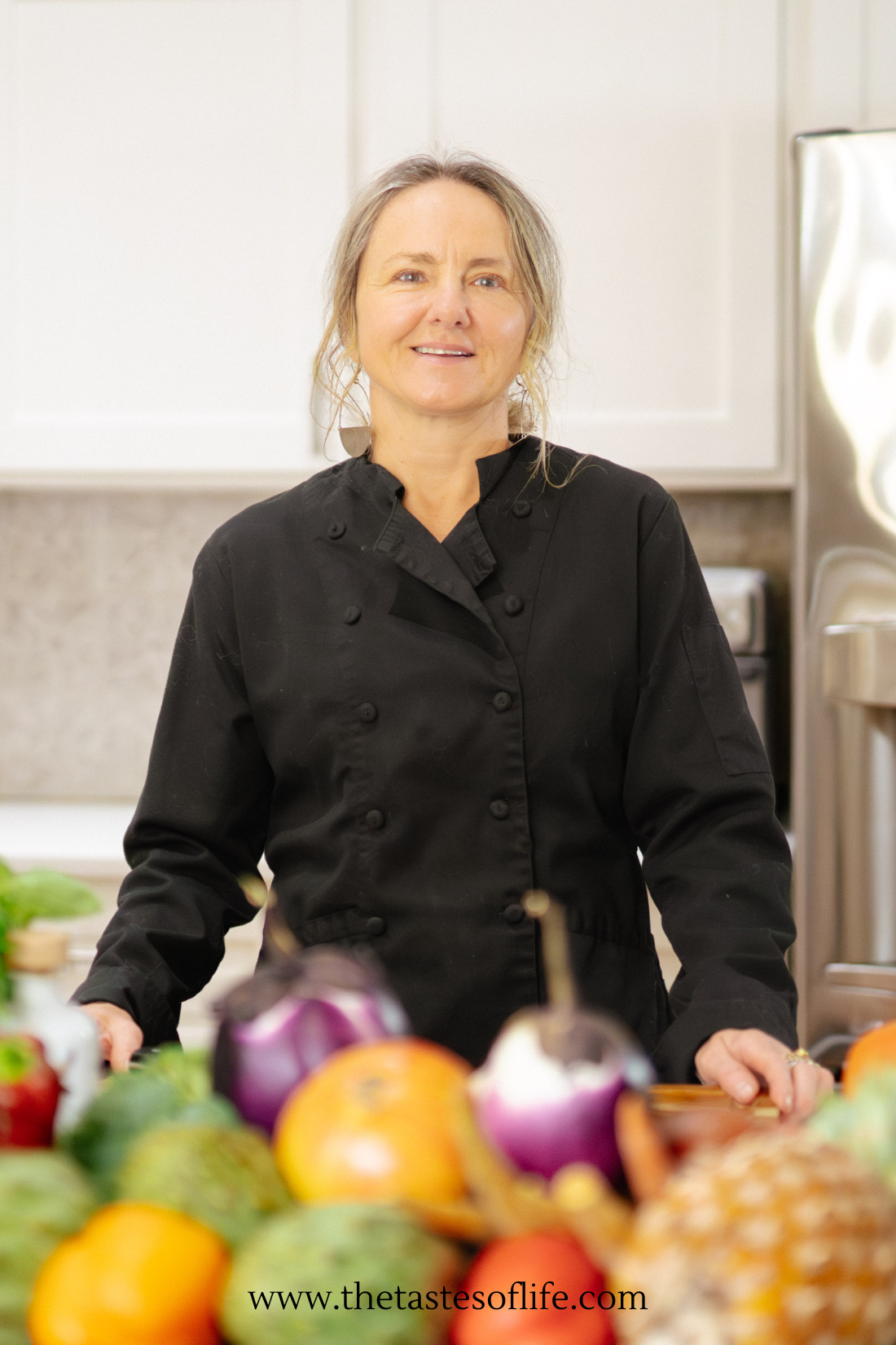 A chef in a black jacket stands in a bright kitchen behind a counter filled with colorful fruits and vegetables.