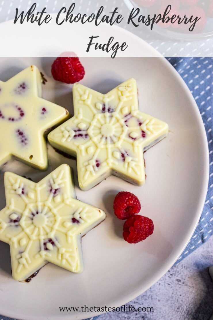 Three white chocolate raspberry fudge pieces shaped like snowflakes are arranged on a white plate with fresh raspberries beside them. The text White Chocolate Raspberry Fudge appears at the top.