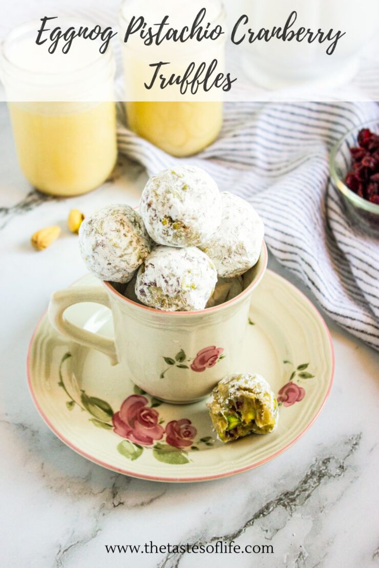 A teacup with a floral design filled with powdered sugar-coated eggnog pistachio cranberry truffles, one with a bite taken out, on a matching saucer with ingredients and a striped cloth in the background.