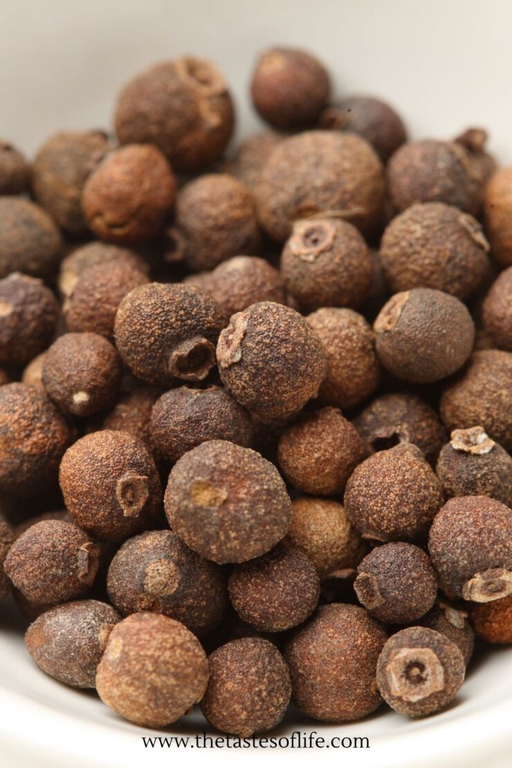 A close-up of whole allspice berries in a white bowl. The round, brown berries have a rough texture, and the image shows them tightly packed together.