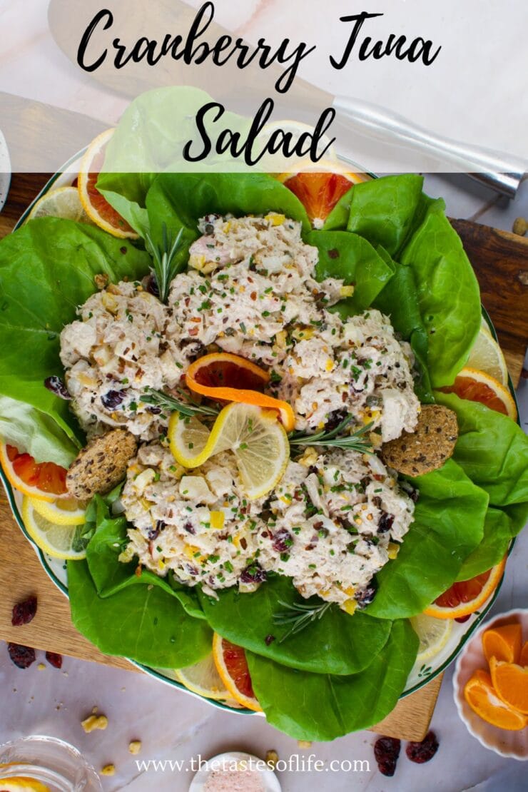 A plate of cranberry tuna salad served on a bed of green lettuce, garnished with lemon slices, orange slices, herbs, and crackers. The dish is colorful and fresh, viewed from above.