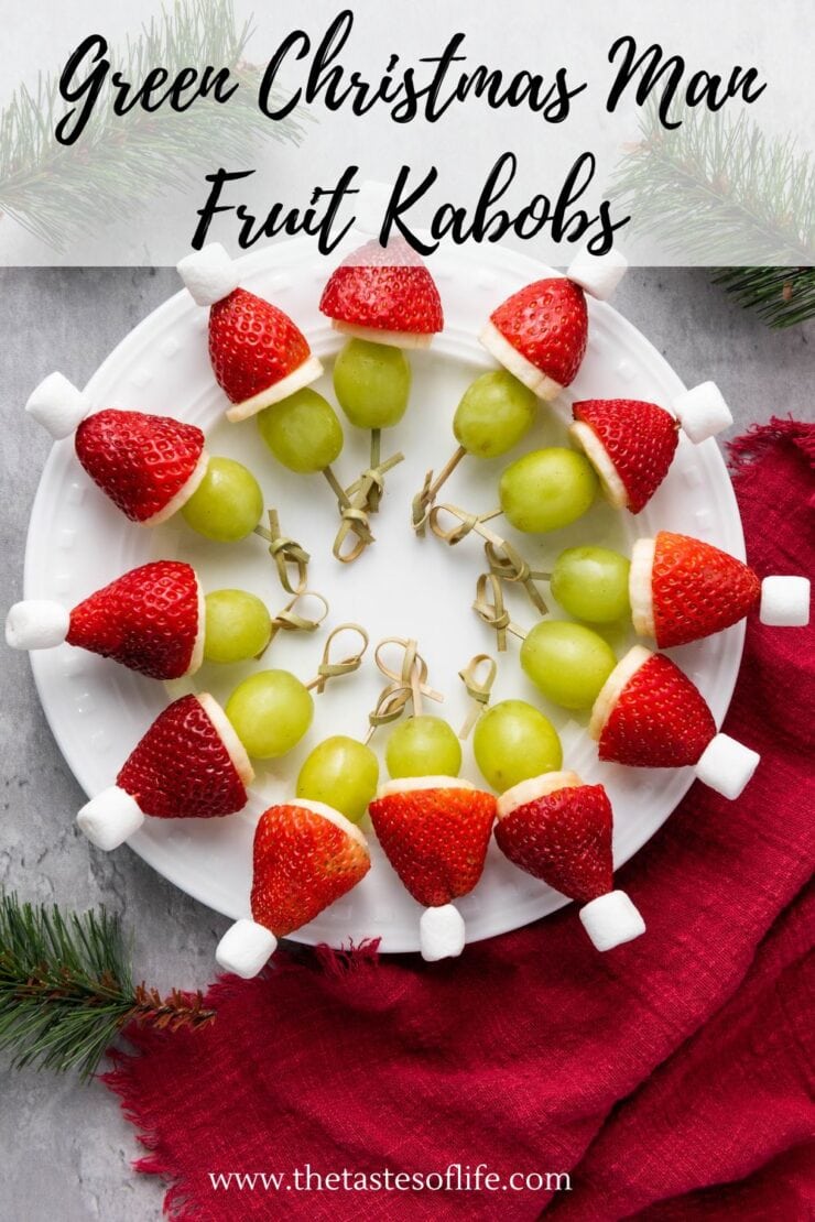 A white plate with fruit kabobs shaped like Christmas hats, made of green grapes, strawberries, and marshmallows, arranged in a circle on a red napkin with pine branches nearby. Text reads Green Christmas Man Fruit Kabobs.