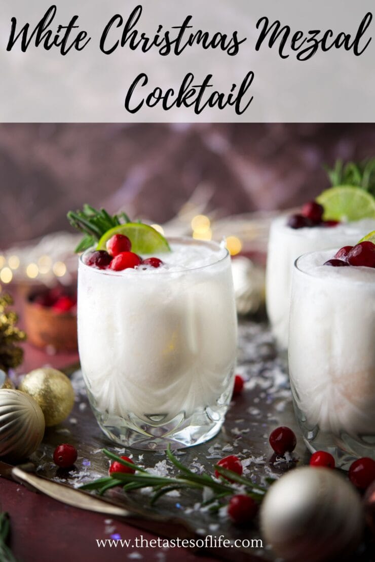 Two frosty white cocktails in clear glasses, garnished with cranberries, rosemary, and a lime slice, sit on a festive tray with ornaments and fairy lights in the background. Text reads &ldquo;White Christmas Mezcal Cocktail.&rdquo;.