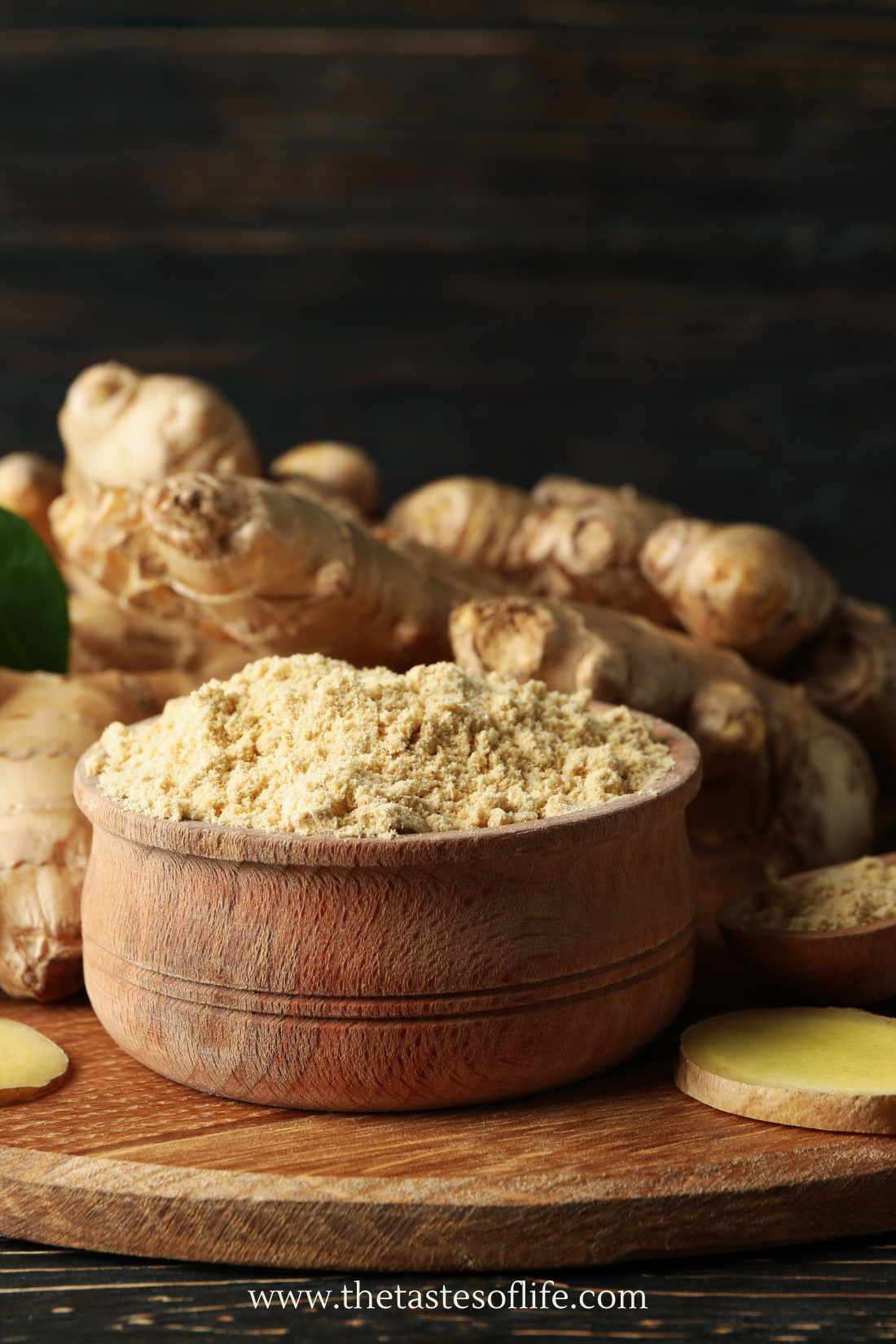 A wooden bowl filled with ground ginger sits on a board, surrounded by fresh ginger roots and ginger slices, with a dark wooden background.