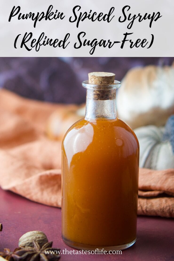 A glass bottle filled with pumpkin spiced syrup sits on a table with autumn-colored cloth, decorative pumpkins, and spices in the background. Text on the image reads Pumpkin Spiced Syrup (Refined Sugar-Free).
