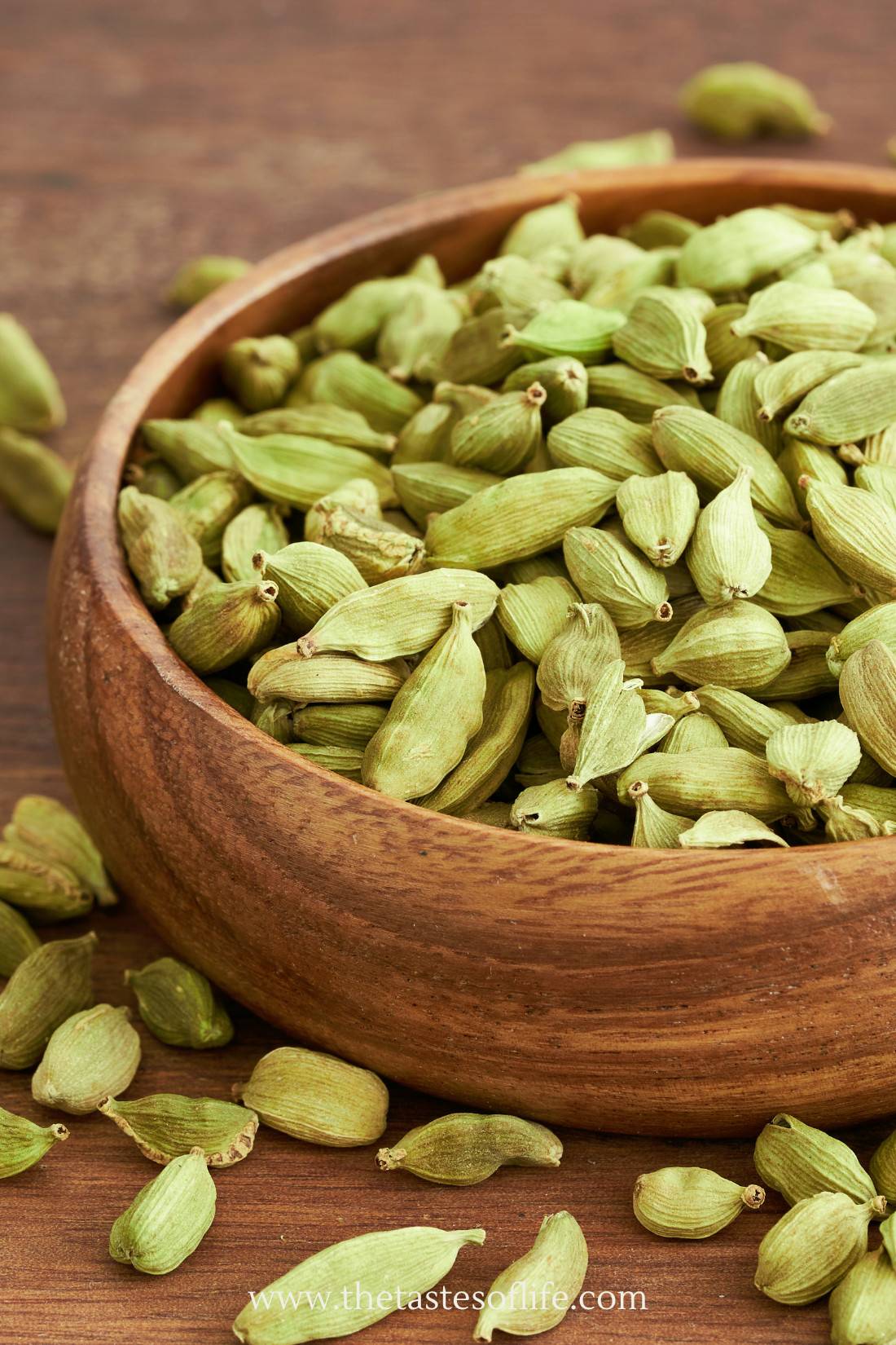 A wooden bowl filled with green cardamom pods sits on a wooden surface, with some pods scattered around the bowl.