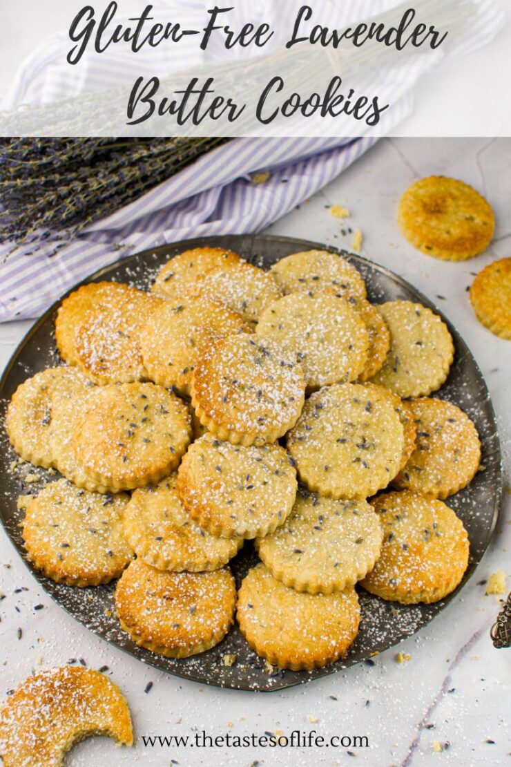 A plate of gluten-free lavender butter cookies is arranged on a marble surface, dusted with powdered sugar. Some lavender sprigs and cookies are scattered around for decoration.