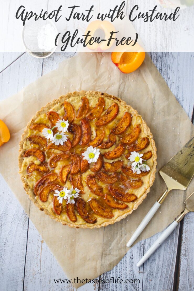 Gluten-free apricot tart with custard, garnished with white flowers, on parchment paper. Sliced apricots and a pie server are nearby on a rustic white wooden surface. Text over the image reads Apricot Tart with Custard (Gluten-Free).
