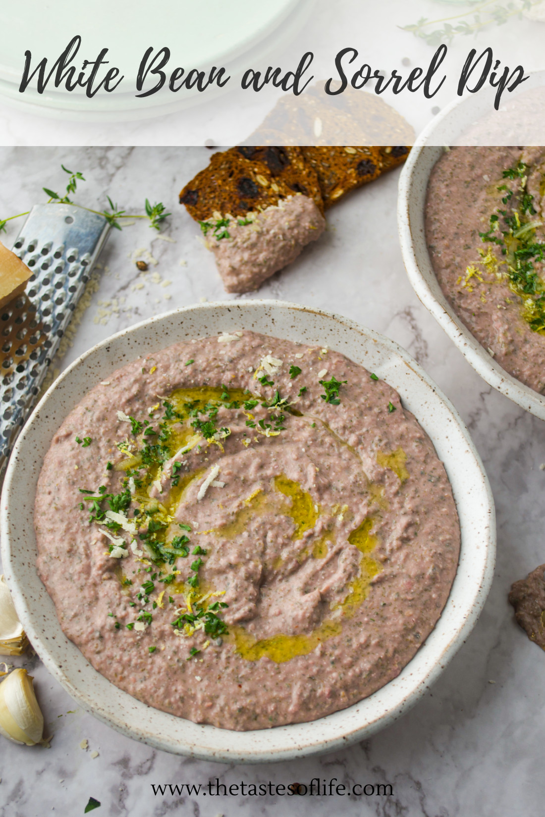 A bowl of creamy white bean and sorrel dip garnished with herbs and olive oil, with crackers, garlic, and a grater nearby on a light surface. Text: White Bean and Sorrel Dip and thetasteoflife.com.