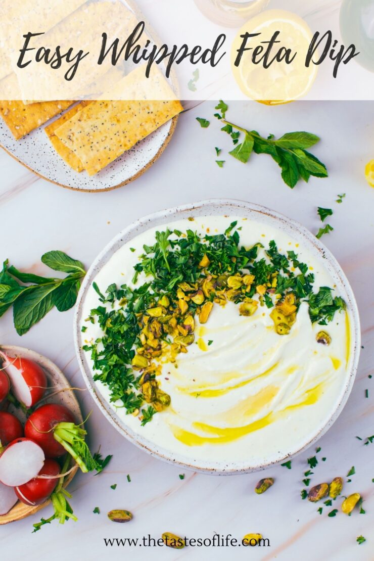A bowl of whipped feta dip topped with herbs and pistachios, drizzled with olive oil, next to crackers, fresh mint, radishes, and lemon slices on a light surface.