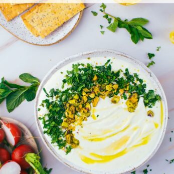 A bowl of whipped feta dip topped with herbs and pistachios, drizzled with olive oil, surrounded by crackers, fresh herbs, radishes, and lemon slices on a marble surface.