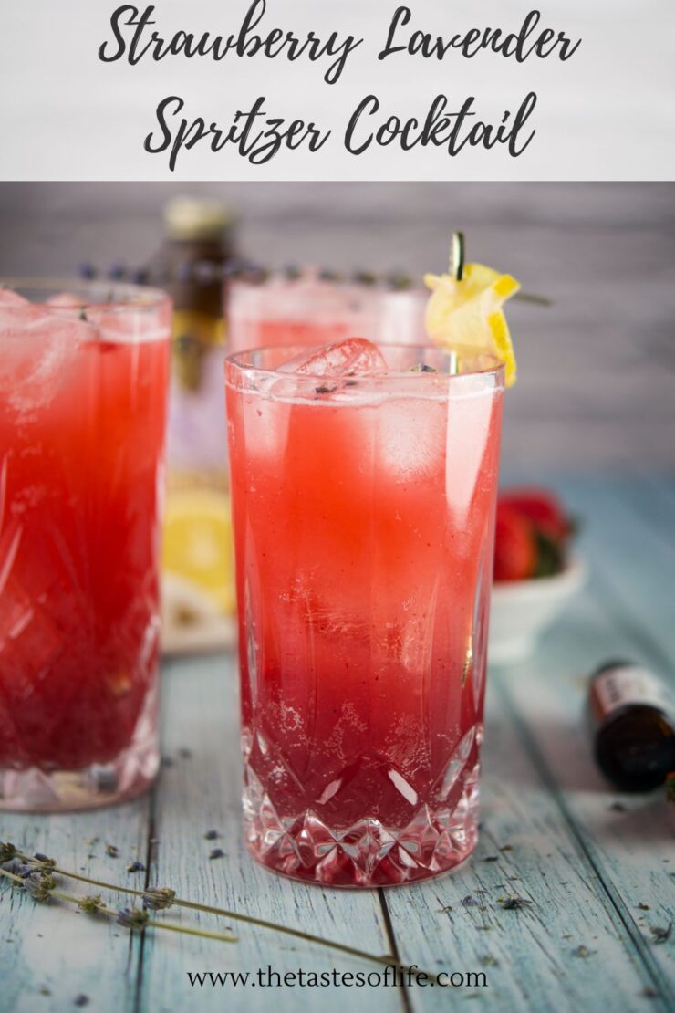 Two tall glasses of strawberry lavender spritzer with ice and lemon garnish sit on a blue wooden surface. Fresh strawberries, a lemon, and a lavender sprig are nearby. Text reads Strawberry Lavender Spritzer Cocktail.